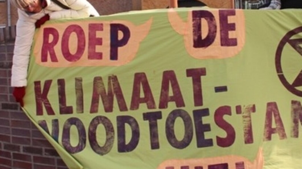 Foto van een actievoerder die buiten het gemeentehuis van Castricum een spandoek ophangt met de tekst 'Roep de klimaatnoodtoestand uit'