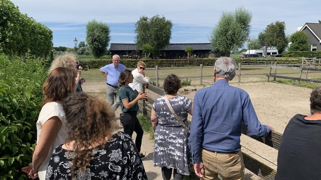 Fractieleden van GroenLinks en PvdA staan bij een zandpaddock van zorgboerderij De Buitenkans. Ze luisteren naar uitleg van een medewerker van De Buitenkans.