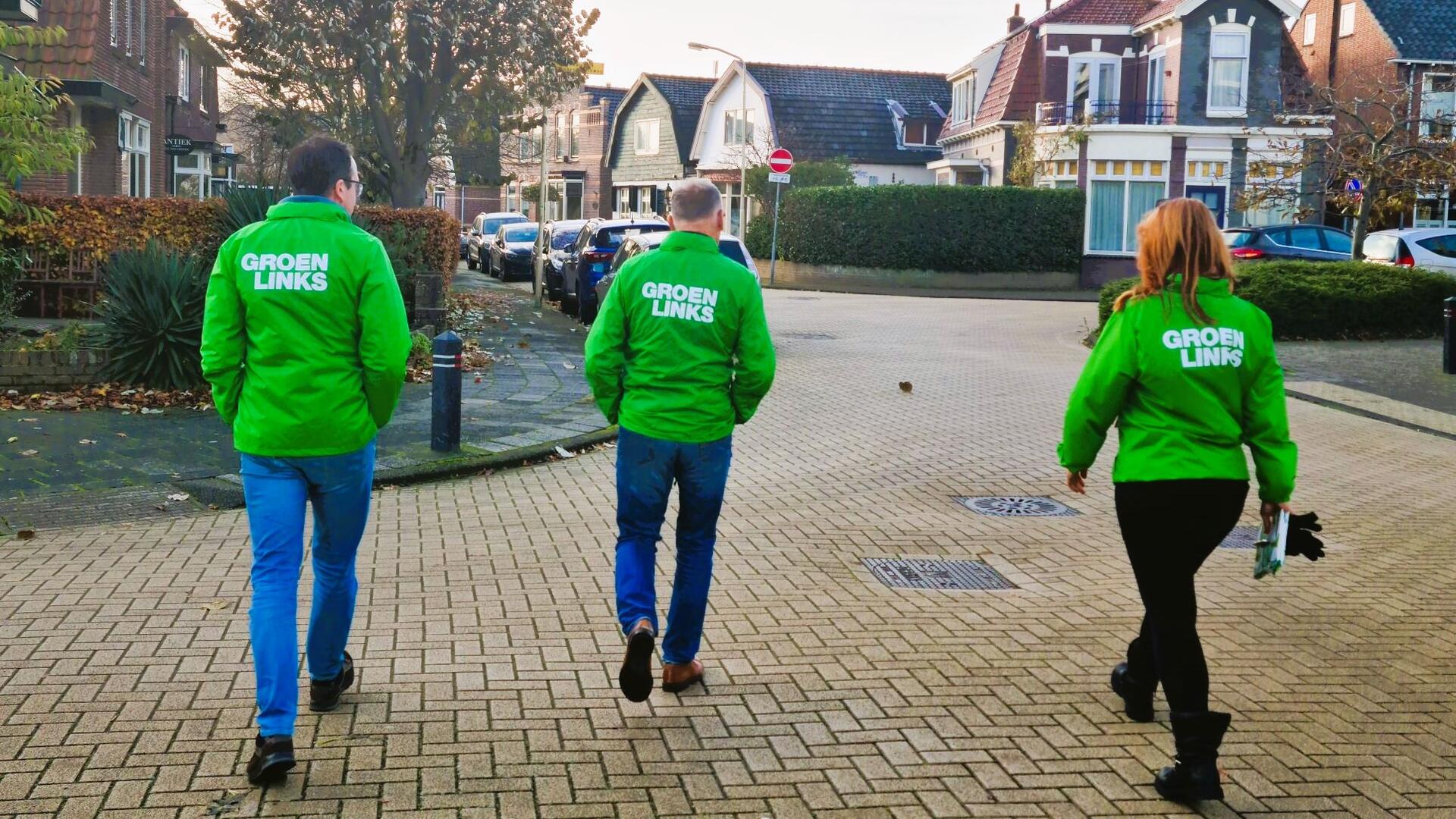 De buurt in: foto van 3 mensen in GroenLinks jasjes, wandelend over straat.
