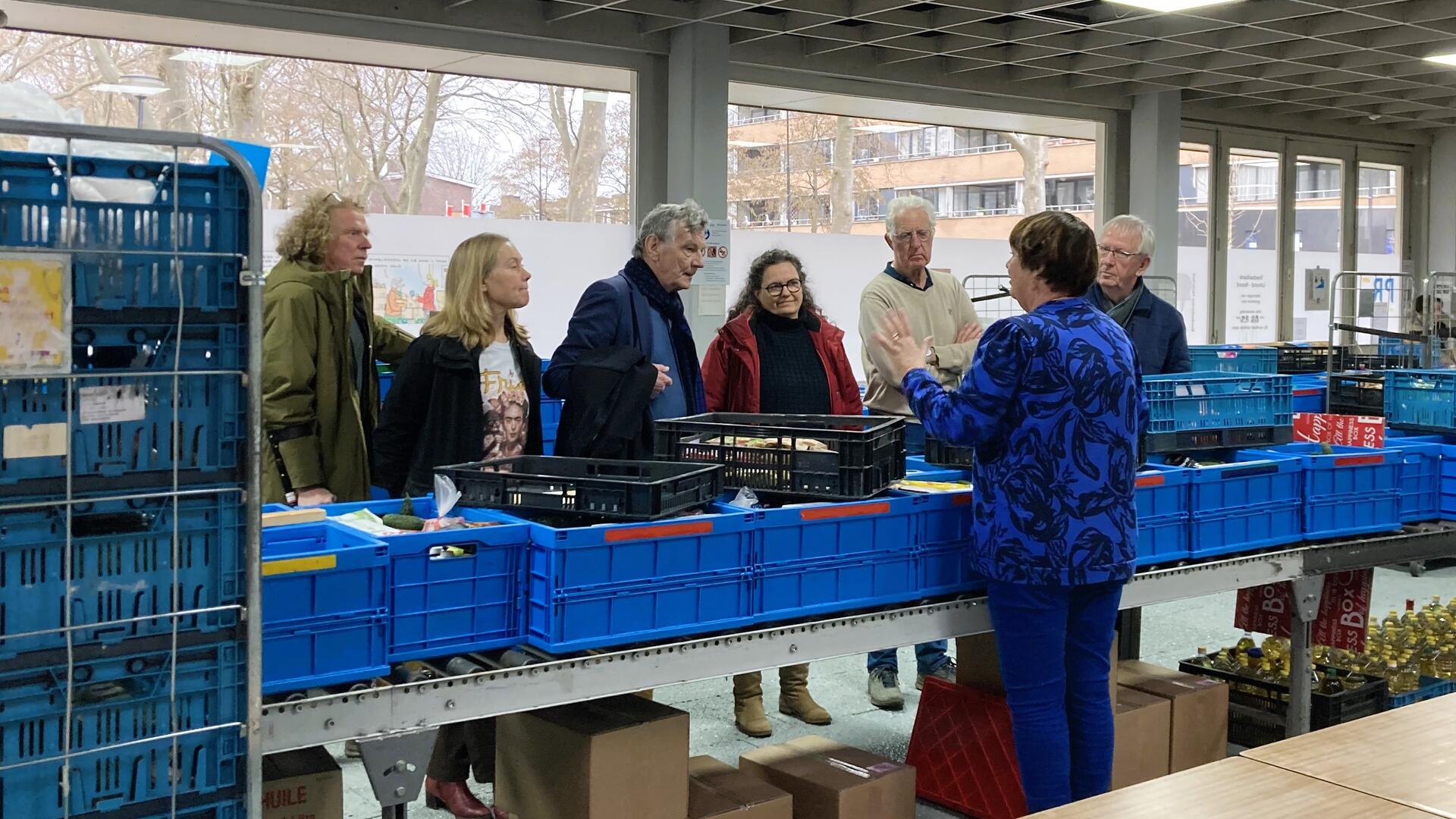 GroenLinksers bezoeken de Voedselbank in IJmond. Ze luisteren naar een vrouw die voor een rij blauwe kratten op een tafel uitleg geeft over de voedselinzameling en verdeling in kratten.