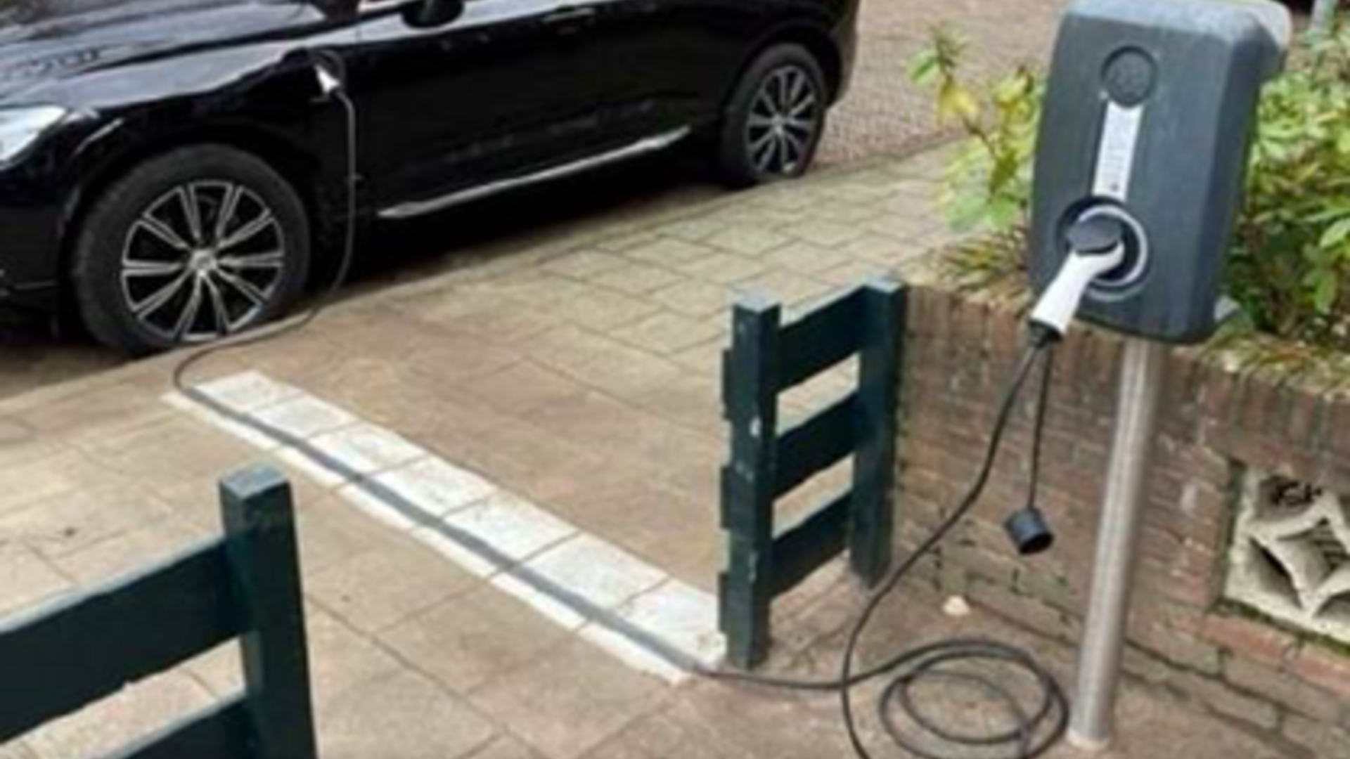 Foto van een laadpaal in een voortuin, een auto geparkeerd naast de stoep en een laadkabel die via kabelgoottegels van de laadpaal naar de auto gaat.