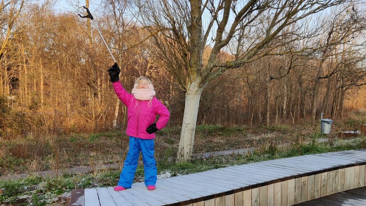 Meisje in roze winterjas en blauwe broek steekt haar plog-stok trots in de lucht, terwijl ze op een bankje staat met het winterse bos op de achtergrond.