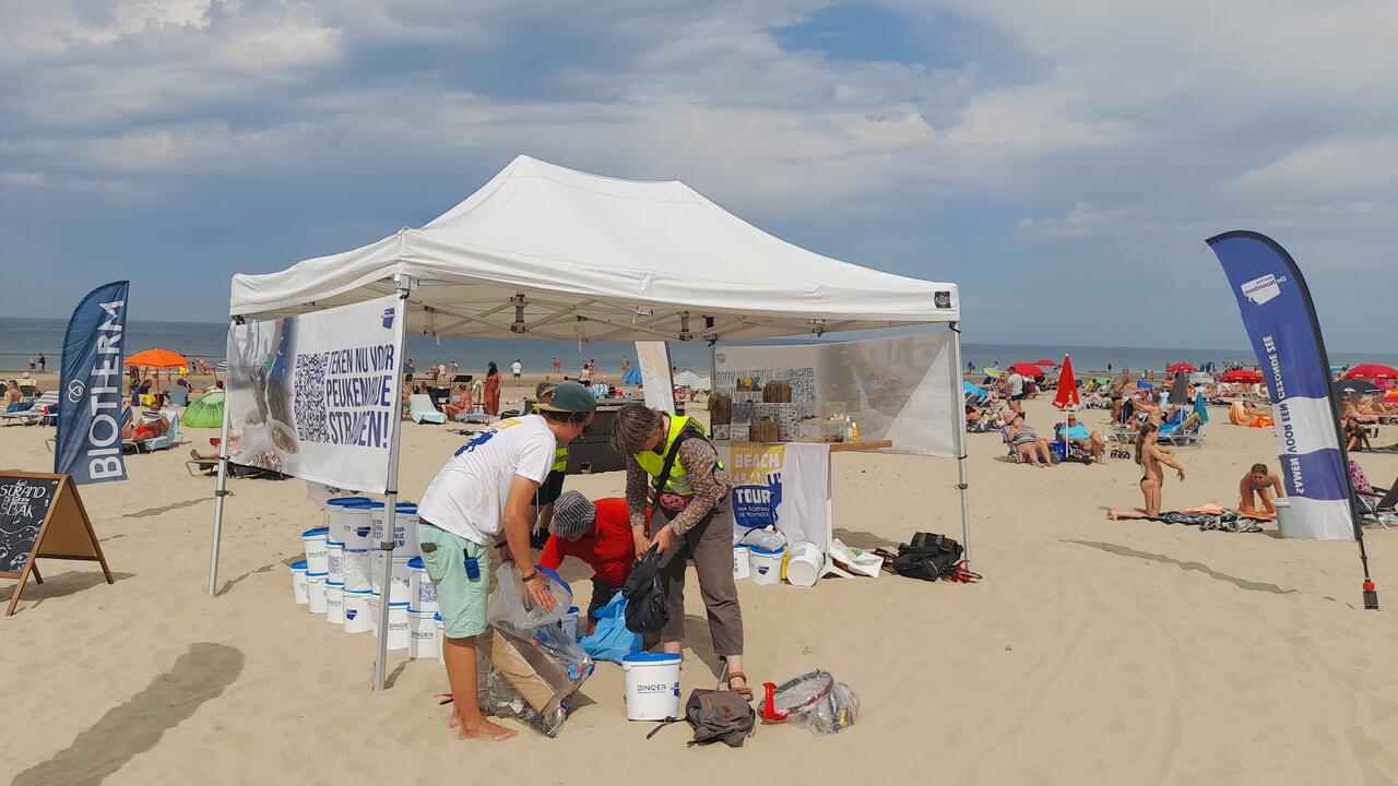 Een witte partytent op het strand, met op de zijkant een banner met de tekst 'Teken nu voor peukenvrije stranden'. Onder en voor de tent zijn vrijwilligers geraapt afval aan het verzamelen. Rond de tent zitten strandgangers.